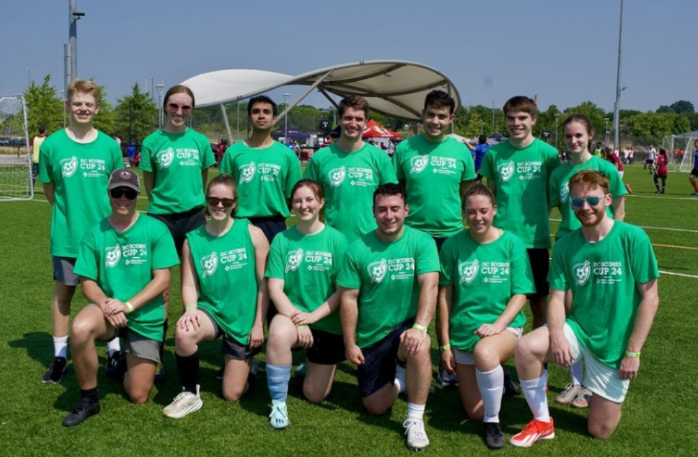 A group of young men and women standing and kneeling together and looking at the camera. They are wearing green shirts labeled with the words, "DC Scores Cup 2024."