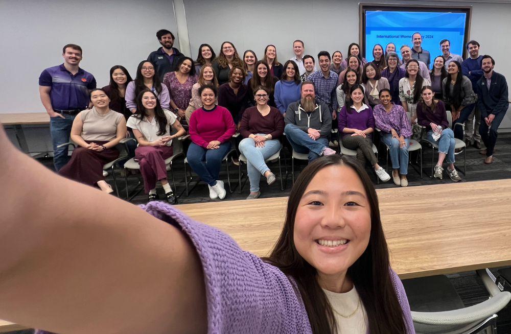 A large group of men and women smiling together, with one woman in front holding the camera selfie-style. Most of them are wearing purple.