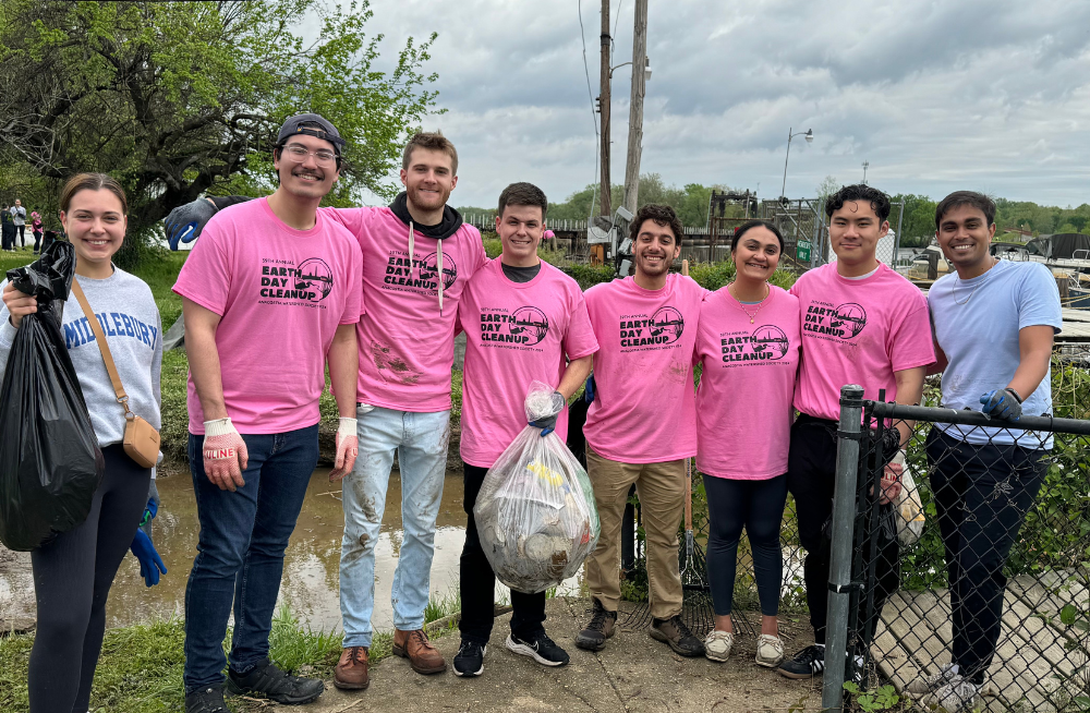 A group of young men and women standing together outside. They are smiling at the camera and a few hold bags of trash. Many wear shirts reading: "Earth Day Cleanup."