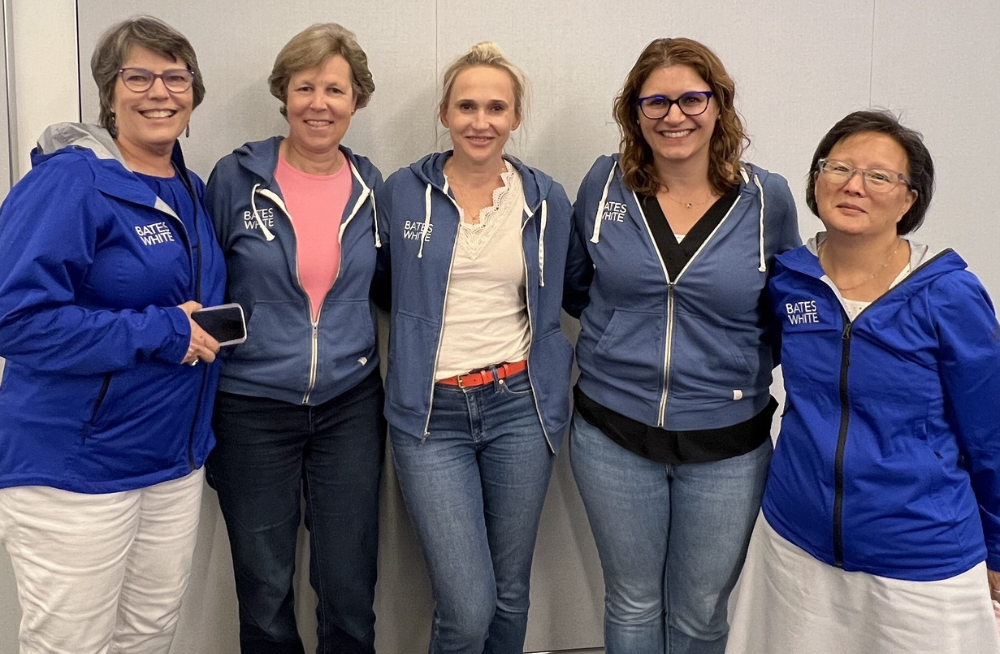 A group of five women, standing arm-in-arm and smiling at the camera. They are wearing blue jackets with the "Bates White" logo on them.