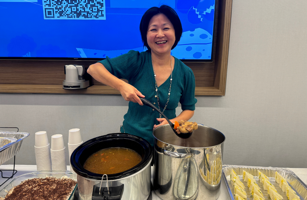 A smiling woman is holding a ladle filled with food over a pot. She stands behind a table clad with food.