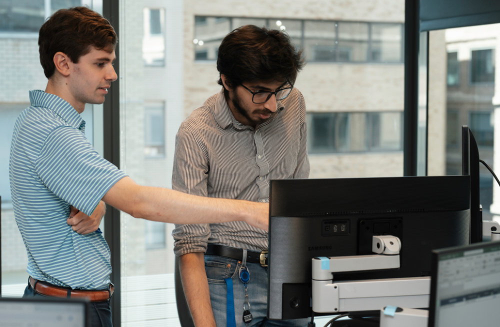 Two young men are gathered around a computer monitor. One of the men is pointing at the monitor.