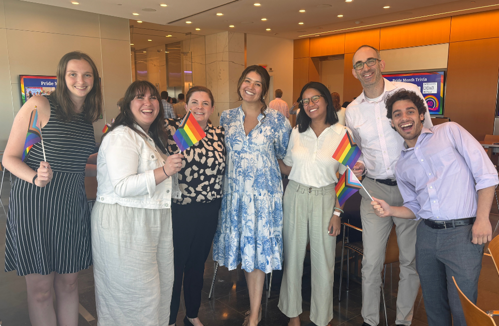 A group of 7 people (5 women, 2 men) stand together, smiling. Several of them are holding rainbow-colored Pride flags.