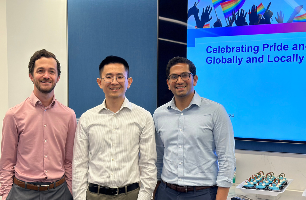 Three men stand together smiling at the camera. A tray of cupcakes is behind them, as well as a large screen reading: "Celebrating Pride and Allyship."