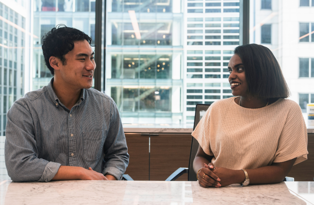 A man and woman sitting beside each other behind a table. The window behind them shows tall buildings.