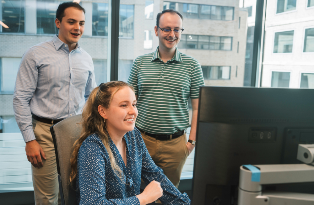 A woman seated in a chair and two men standing behind her are all smiling at a computer monitor.