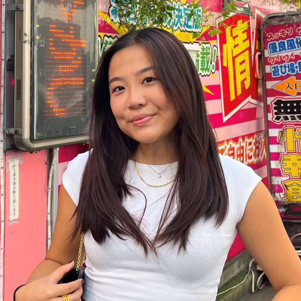 A headshot of a young smiling woman. Behind her are colorful signs and symbols in a foreign language.