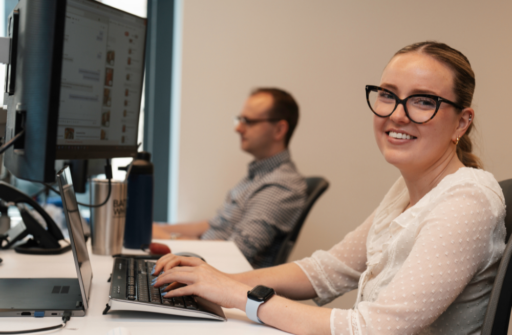 A young woman is seated in front of a computer monitor, typing while smiling at the camera. In the background is a young man staring at his monitor.