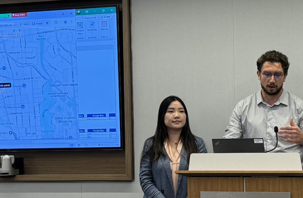 A man is standing behind a lectern and speaking into a microphone. A woman stands beside him. Beside them both is a screen showing a map of Baltimore, Maryland.