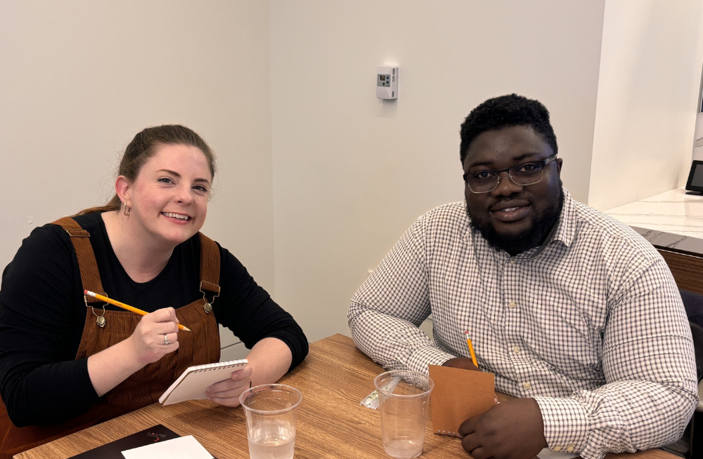 A young man and woman are seated at a table, each holding a pencil and a notepad. Both are smiling at the camera.