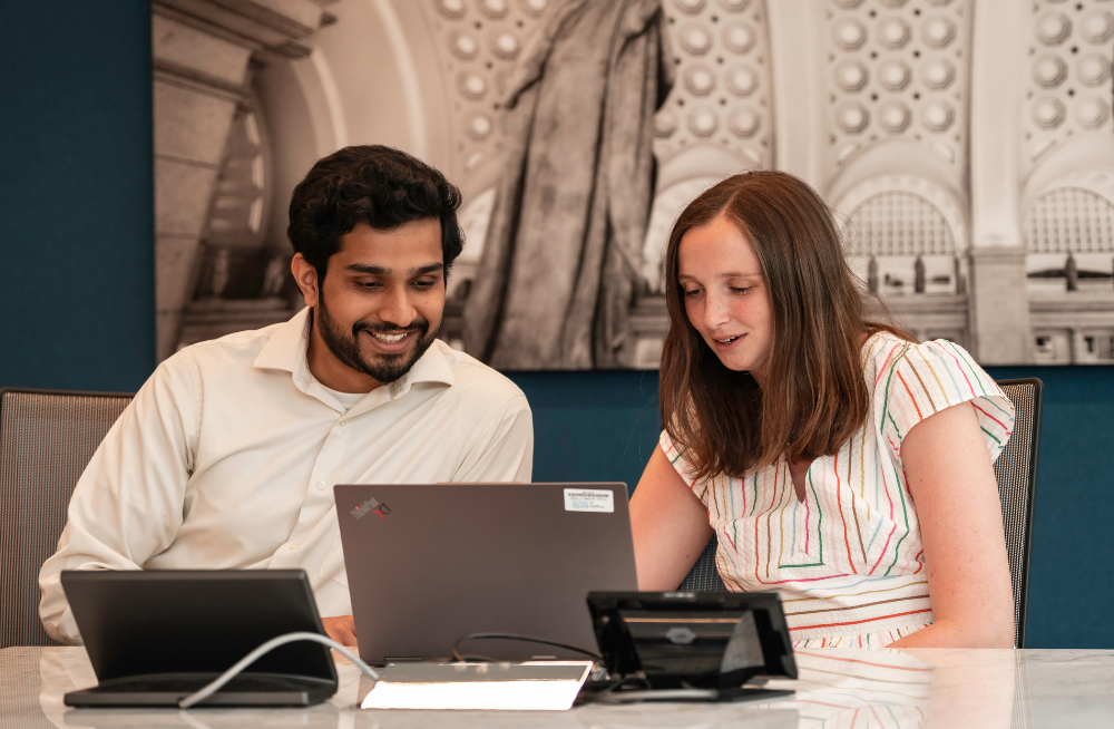 A young man and woman sit at a desk, staring at a laptop screen.