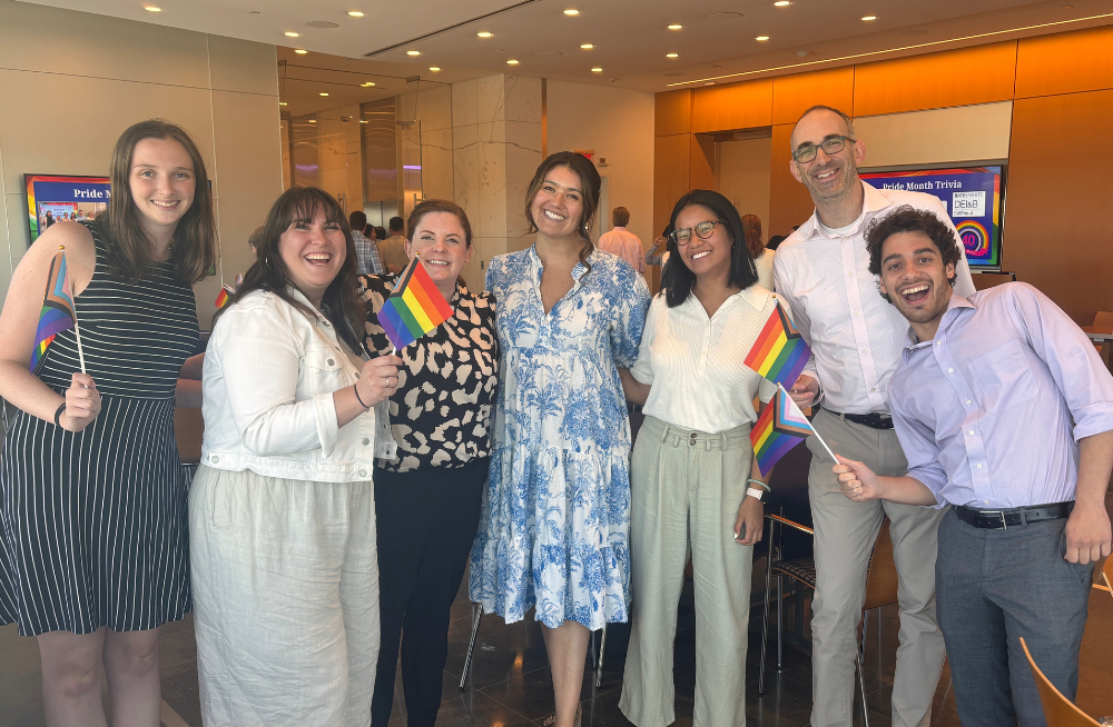 A group of men and women stand together smiling at the camera; several of them are holding rainbow-colored Pride flags.