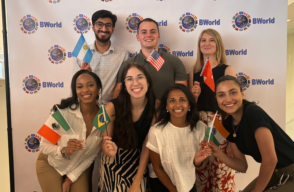 A group of men and women standing together, smiling at the camera. A backdrop behind them is peppered with the word "BWorld." They are holding flags from various nations.